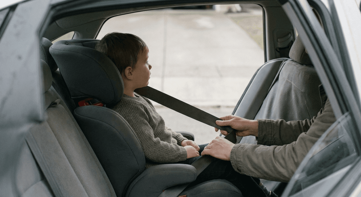Child in a high-back booster seat while a parent pulls the seat belt across the lap