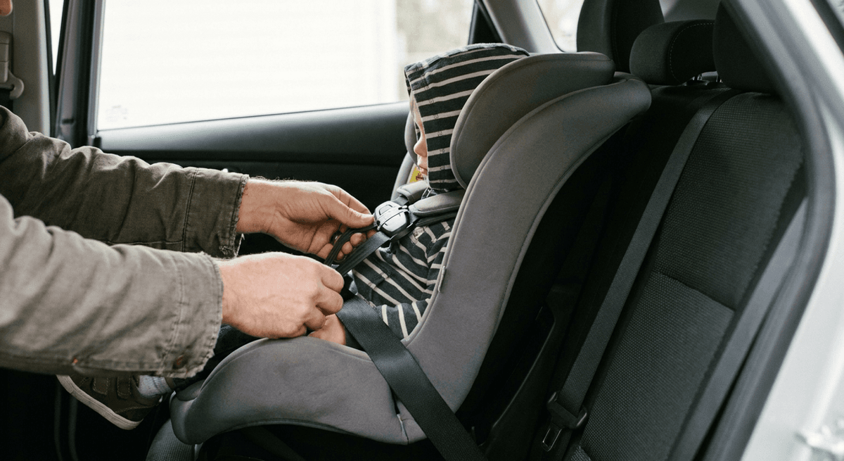 Parent hands tightening the harness strap on a toddler in a grey rear-facing car seat in a sedan
