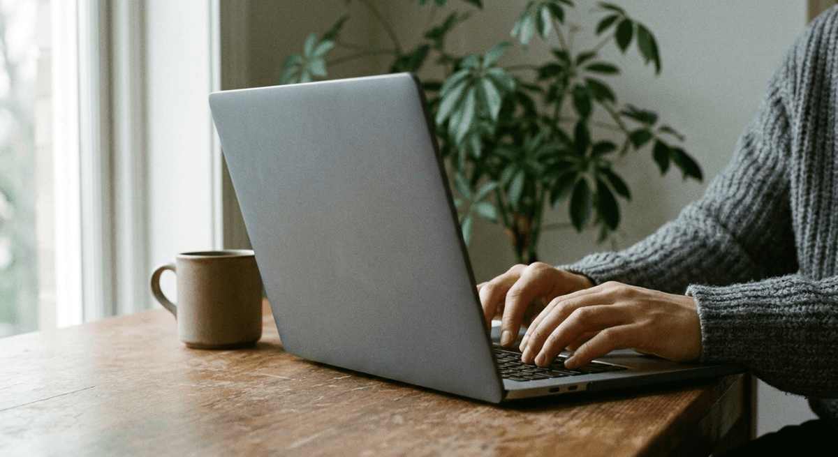 Hands typing on a laptop seen from behind its plain grey lid, with a mug and a plant on a wooden desk