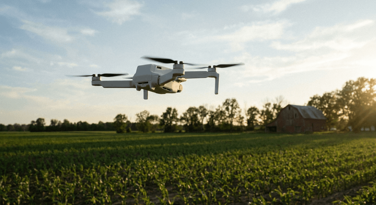 A small white drone hovers above a green farm field with a red barn at sunset