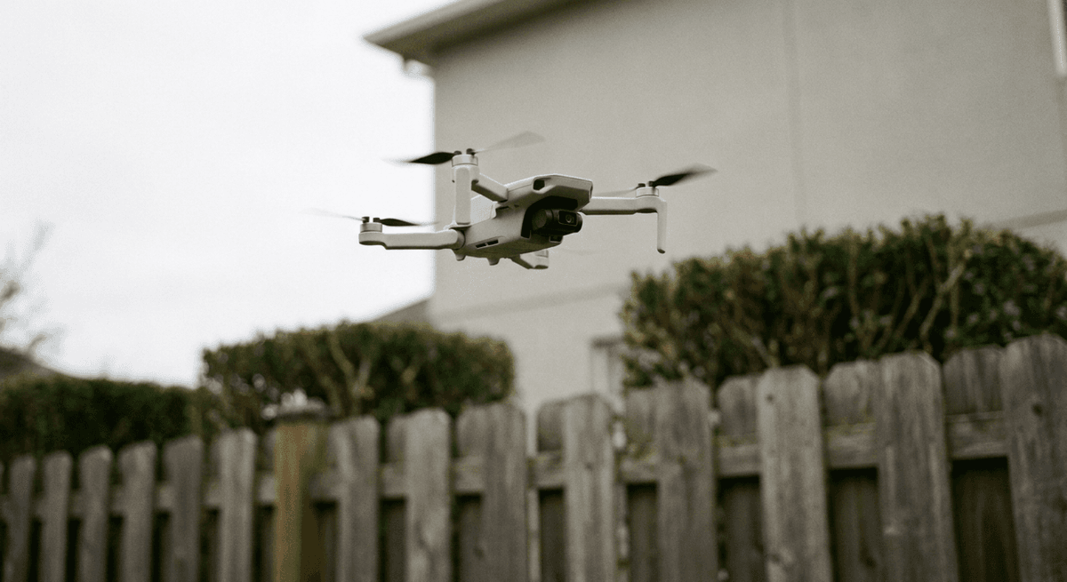 Small white drone hovering above a wooden backyard fence and hedge