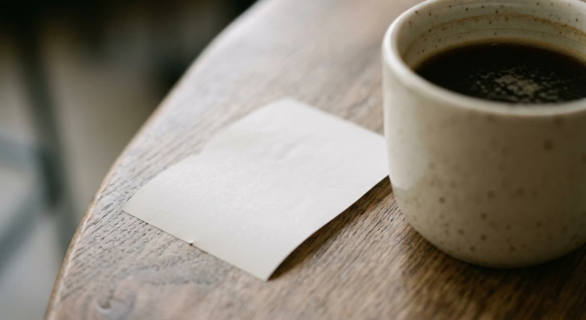 a close crop of a direct-deposit-style bank receipt beside a coffee cup, figures blurred, morning light