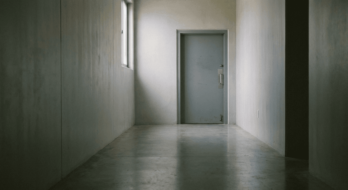 un pasillo de concreto vacío con poca luz que lleva a una sola puerta de acero gris cerrada