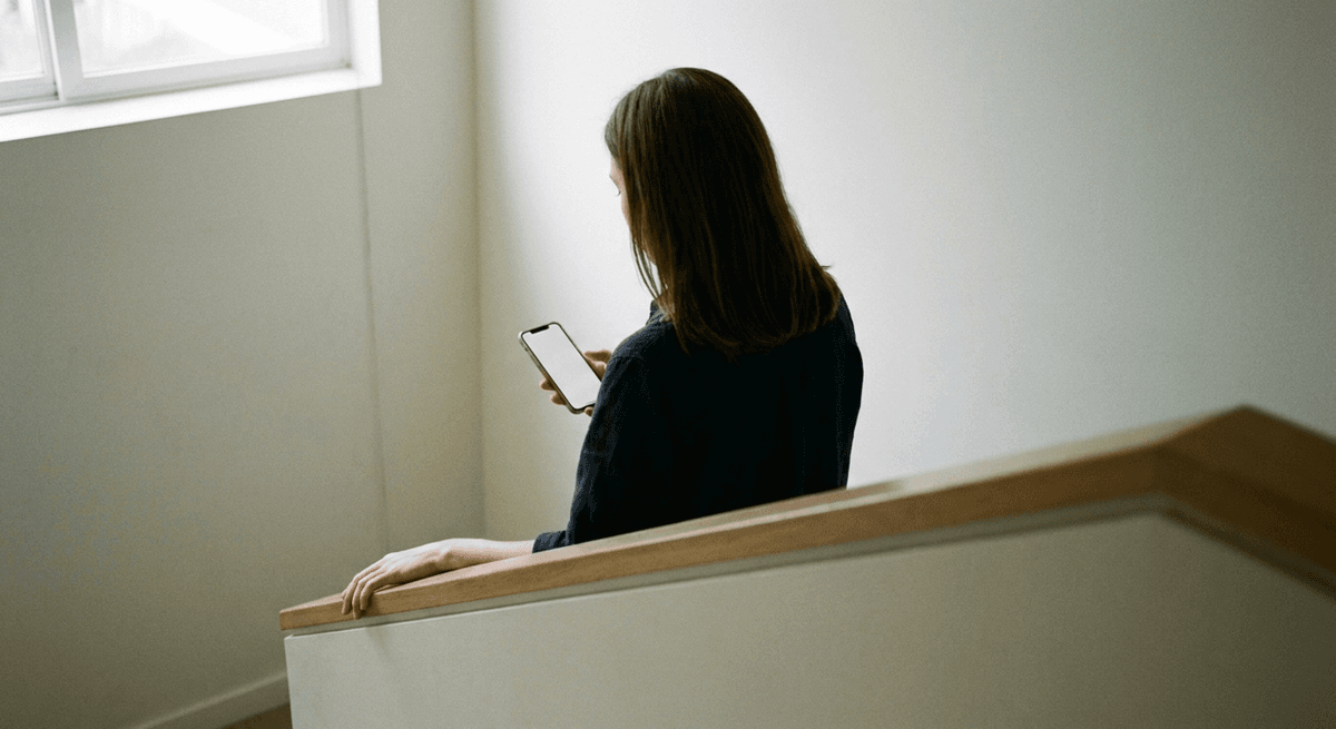 A woman seen from behind on a stairwell landing looking down at a smartphone, one hand resting on the handrail