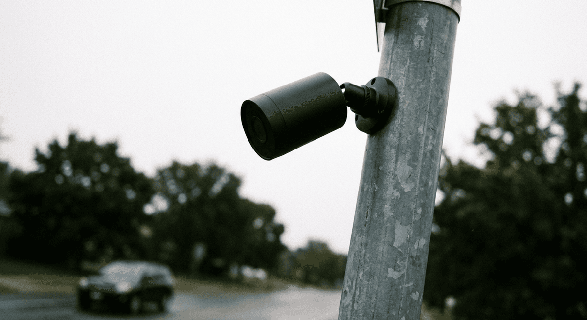 Plain black bullet security camera mounted on a metal pole above a blurred street