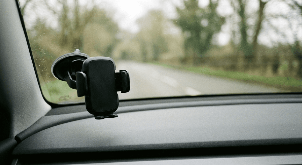 A black suction-cup phone cradle stuck low on a car windshield with a quiet tree-lined road beyond the glass